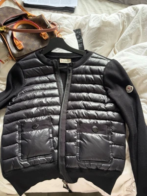 Svart cardiganjacka från Moncler - Snygg svart cardiganjacka från Moncler med quiltad framsida och två stora fickor med knapp. Ärmar och rygg i mjuk stickad kvalitet, framstycke i glansigt material. Dragkedja framtill och Moncler-logga på ärmen. Perfekt för lager på lager och streetstyle. OBS har ej äkthetsbevis!