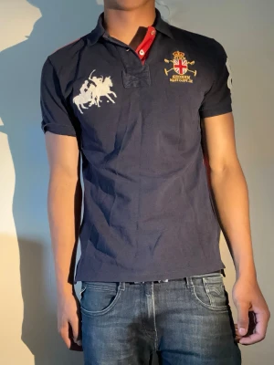 Ralph lauren piké - Marinblå pikétröja från ralph lauren med broderad logga och polospelare på bröstet. Tröjan har korta ärmar, krage med röda detaljer och knappar framtill. Perfekt för en sportig och stilren look.