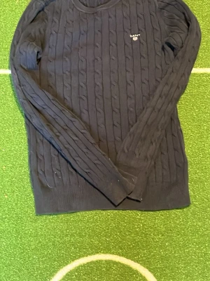 Mörkblå stickad tröja Gant - Kabelstickad mörkblå tröja från Gant med rund halsringning och diskret logga broderad på bröstet. Tröjan har långa ärmar och ribbade muddar vid ärmslut och nederkant. Perfekt för en klassisk och stilren look. Inga fläckar.