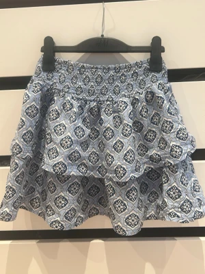 Blå mönstrad kortkjol från Gina Tricot - Supersöt blå kortkjol från Gina Tricot med vitt och mörkblått grafiskt mönster. Kjolen har resår i midjan och volangdetaljer som ger en luftig och lekfull känsla. Perfekt för dig som gillar en avslappnad men trendig stil.