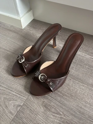 Bruna slip-in pumps med silverdetalj - Snygga bruna slip-in pumps med öppen tå och hög smal klack. Skorna har en dekorativ silverfärgad spänne framtill och kontrastsömmar längs kanterna. Tillverkade i glansigt läderliknande material för en stilren look. Köpt secondhand, men ser oanvända ut. 