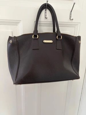 Mörkbrun handväska från Jimmy Choo - Snygg mörkbrun handväska från Jimmy Choo i äkta läder med guldfärgade metalldetaljer och dubbla handtag. Väskan har en klassisk, strukturerad form och stängs med dragkedja. Perfekt för dig som gillar lyxiga accessoarer med stilren design.den här viss defekter som ni kan se men går nog att fixa 