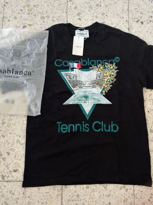 Svart Casablanca Tennis Club t-shirt - Casablanca Tennis Club t-shirt i svart med färgglad grafik framtill och tryckt logga. Klassisk rund hals och korta ärmar. Tillverkad i mjuk bomull för skön känsla. Perfekt för dig som gillar streetwear och sportig stil.