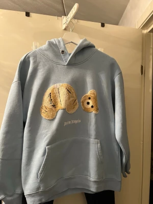 Ljusblå hoodie från Palm Angels - Ljusblå hoodie från Palm Angels med broderad nallebjörn på framsidan. Hoodien har stor magficka, huva och avslappnad passform. Perfekt för dig som gillar streetwear och vill sticka ut med en unik design.