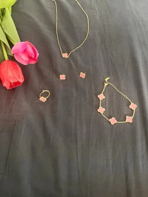 Smyckesset med rosa fyrklöver - Snyggt smyckesset med halsband, armband, ring och örhängen i guldfärgad metall. Alla delar har en rosa fyrklöverformad detalj med guldfärgad kant. Perfekt för dig som gillar söta och färgglada accessoarer. Det är stainless steel