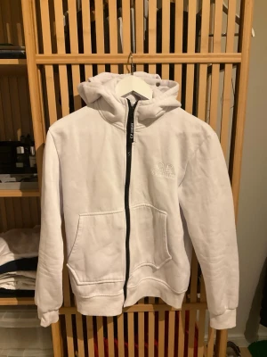 Vit hoodie från C.P. Company - Snygg vit hoodie från C.P. Company med dragkedja framtill och broderad logga på bröstet. Hoodien har en klassisk huva och stora fickor framtill. Tillverkad i mjukt material som känns skönt mot huden. Perfekt för en clean och stilren look. Storlek XS-S.