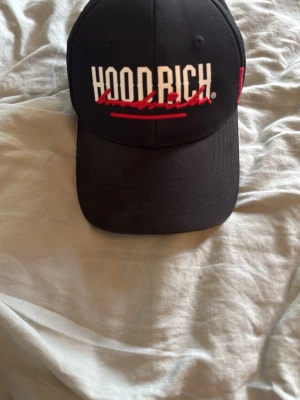 Svart Hoodrich keps med brodyr - Cool svart keps från Hoodrich med vit och röd brodyr på fronten och sidan. Kepsen har böjd skärm och detaljer som streetwear-etikett på sidan. Materialet är bomull och designen är klassisk med snapback-justering. Perfekt för dig som gillar urban stil.