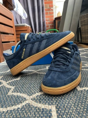 Adidas Handball Spezial navy blue - Mörkblå Adidas Handball Spezial sneakers i mocka med klassiska tre ränder på sidan och gulddetaljer. Skorna har en brun gummisula och låg profil. Perfekta för dig som gillar retrostil och streetwear.