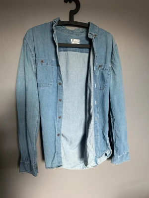Ljusblå denimskjorta från Cubus - Säljer en ljusblå denimskjorta från Cubus med klassisk krage, två bröstfickor och knappar framtill. Skjortan är långärmad och har en avslappnad passform som är perfekt för en cool och enkel look.