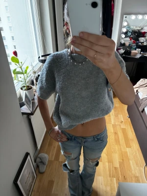 Grå stickad topp  - En grå, stickad topp med korta ärmar och pärlknappar i ryggen eller där fram Mjuk och fluffig känsla, perfekt för att skapa en chill och trendig look. Enkel att matcha med jeans eller kjol för en avslappnad stil.