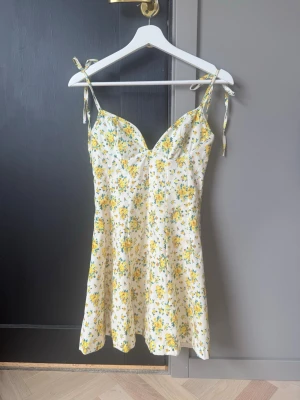 Blommig klänning - Söt kortklänning med blommigt mönster i gult och grönt på vit botten. Klänningen har v-ringad front, smala axelband med knyt och en lätt utsvängd form. Perfekt för sommarens dagar och ger en fräsch och ungdomlig look.💛💚