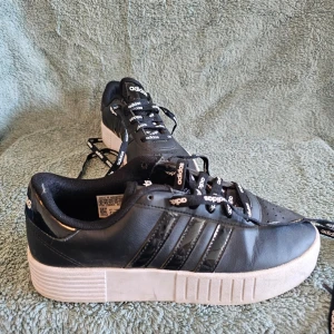 Svarta Adidas sneakers med vita sulor - Snygga svarta Adidas sneakers med klassiska tre ränder på sidan och vit tjock sula. Skorna har svarta snören med Adidas-logga och är tillverkade i läder och syntetmaterial. Perfekt för dig som vill ha en stilren och sportig look.