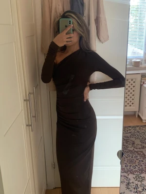 Brun långklänning med oneshoulder - Brun långklänning med oneshoulder-design och lång ärm. Klänningen har en figurnära passform som framhäver siluetten och är perfekt för dig som vill ha en elegant och stilren look. Tillverkad i ett mjukt och stretchigt material.