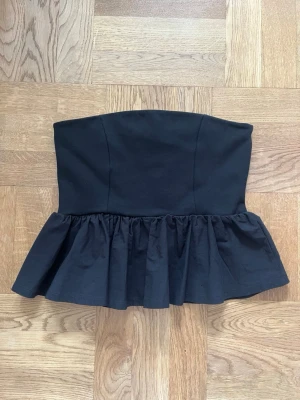 Svart tube topp med volangkant - Svart tube topp med volangkant nedtill. Modellen är croppad och har en bandeauformad överdel. Storlek S. Köpte den på Kappahl för några år sedan men har aldrig använt den.