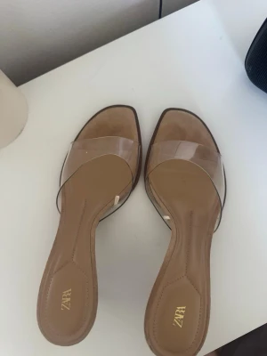 Transparenta klackar från Zara - Säljer ett par trendiga klackar från Zara med transparent rem i plast och beige innersula. Skorna har öppen tå, fyrkantig framdel och smal klack. Används fåtal gånger!