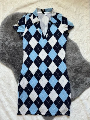 Blå och vit rutig kortklänning - Kortklänning med klassiskt rutigt mönster i ljusblått, vitt och mörkblått. Klänningen har krage, v-ringning och korta ärmar. Materialet känns mjukt och stretchigt, perfekt för en trendig och avslappnad look.