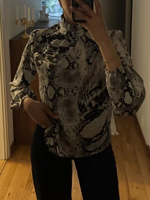 mönstrad blus med hög krage - Snygg blus med svartvitt /grå ormmönster och hög krage. Blusen har långa ärmar med lätt puff och resår vid ärmslut. Passar till både jeans, kostym och kjol. Kolla sista sida för att se baksidan! 
