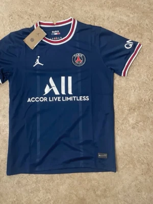 PSG matchtröja blå Jordan x Nike - Snygg blå PSG matchtröja med vita och röda detaljer på krage och ärmslut. Framtill finns PSG-logga, Jordan-logga och sponsortryck. Tillverkad i lätt och ventilerande material, perfekt för fotboll eller träning.