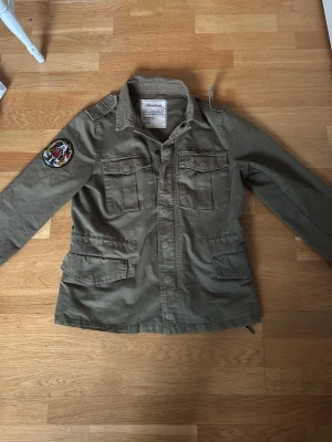 Zadig & Voltaire field jacket - Snygg olivgrön vindjacka från Z&V med fyra framfickor, axelklaffar och ett färgglatt tygmärke på ena ärmen. Jackan har dragkedja och knappar framtill samt justerbar midja. Tillverkad i bomullsmix för en avslappnad look. Saknar knappar på armarna och på axlarna