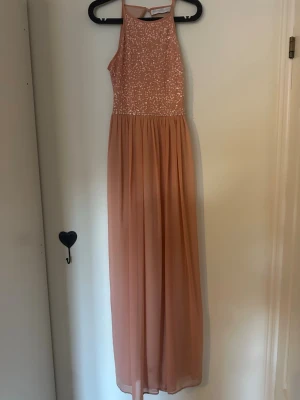 Lång persikofärgad paljettklänning - Elegant långklänning i persikofärg från Vesper Couture. Överdelen är täckt av glittriga paljetter och har en hög halsringning. Kjolen är i ett mjukt, flytande tyg som ger en romantisk känsla. Perfekt för bal eller festliga tillfällen.