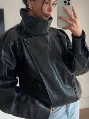 Svart oversized skinnjacka - Cool svart skinnjacka med hög krage och asymmetrisk knäppning. Jackan har en oversized passform, markerade axlar och detaljerade sömmar. Perfekt för dig som vill ha en edgy och trendig look.
