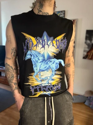 Svart ärmlös topp med blå hästmotiv - Cool svart ärmlös topp med ett stort tryck av en blå häst och grafiska detaljer i gult och lila. Toppen har en rak passform och är gjord i mjukt bomullsmaterial. Perfekt för dig som gillar unika och edgy prints.