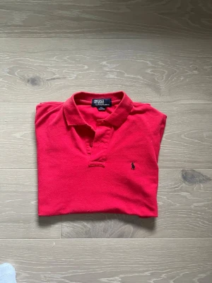 Röd polo från Polo Ralph Lauren - Klassisk röd polo från Polo Ralph Lauren i 100% bomull. Topp med krage, knappar och den ikoniska broderade logon på bröstet. Perfekt för dig som gillar en tidlös och sportig stil.