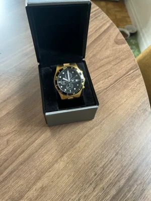 Emporio Armani guld klocka - Snygg guld klocka från Emporio Armani med svart urtavla och flera små visare. Klockan har en robust design och levereras i originalbox med certifikat och manual. Perfekt för dig som vill ha en exklusiv accessoar med stilren look.
