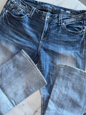 Miss me blå jeans med paljetter - Snygga blå jeans från Miss me med slitningar och coola paljettdetaljer på bakfickorna. Klassisk femficksmodell med kontrastsömmar och låg midja. Perfekt för dig som vill sticka ut med en unik look.