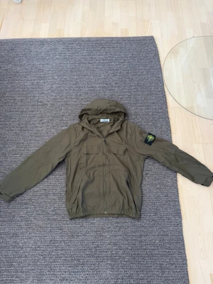 Vindjacka stone Island - Tunn skaljacka. Storlek L. Passar S/M. Kolla måtten på bilderna! Snygg olivgrön vindjacka från Stone Island med huva och dragkedja framtill. Jackan har två bröstfickor med lock, två sidofickor och den klassiska Stone Island-loggan på vänster ärm. Materialet är lätt och perfekt för blåsiga dagar.