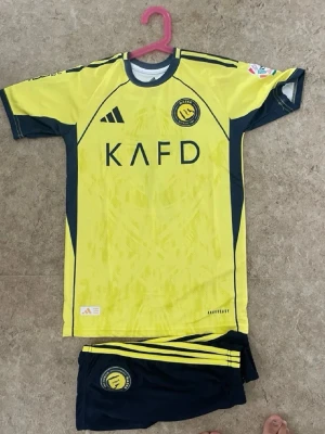 Gul och svart Al Nassr fotbollströja - Säljer en gul och svart Al Nassr fotbollströja med matchande shorts. Tröjan har tryck med KAFD och klubbens emblem på framsidan, samt Ronaldo och nummer 7 på ryggen. Materialet är lätt och andas, perfekt för träning eller match. Shorts med klubbens logga och gula detaljer.