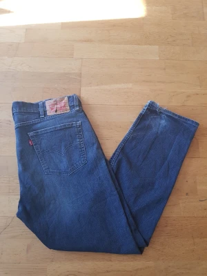 Levi's blå raka jeans - Säljer ett par klassiska blå jeans från Levi's med rak passform och femficksdesign. Jeansen har en ikonisk läderpatch bak i midjan och tydliga kontrastsömmar. Materialet är slitstark denim och färgen är mörkblå. Perfekt för dig som gillar tidlös stil.
