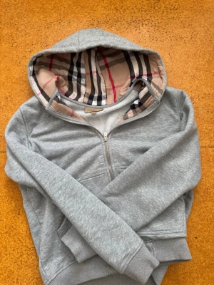Grå hoodie från Burberry med dragkedja - Säljer en grå hoodie från Burberry med klassiskt rutigt foder i huvan. Hoodien har dragkedja framtill, ribbade muddar och är tillverkad i mjukt material. Perfekt för dig som gillar stilrena plagg med exklusiva detaljer.