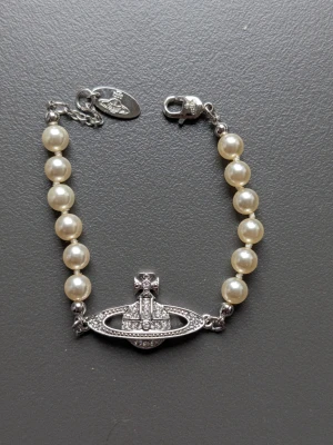 Vivienne Westwood pärlarmband - Snyggt armband från Vivienne Westwood med vita pärlor och ikoniskt silverfärgat Orb-hänge dekorerat med gnistrande stenar. Armbandet har en elegant kedja och lås, och kommer med original ask och tygpåse.