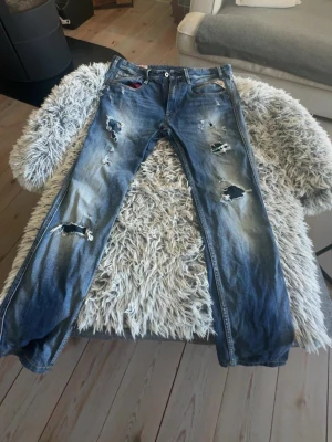Blå slitna jeans med hål - Säljer ett par blå jeans med kraftiga slitningar och hål på flera ställen. Jeansen har en klassisk femficksmodell och raka ben. Perfekta för dig som gillar en edgy och avslappnad stil. Materialet är denim i bomull.