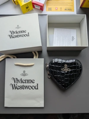 Svart hjärtformad plånbok Vivienne Westwood - Cool svart plånbok från Vivienne Westwood i hjärtform med croco-mönster och silverfärgad logga på framsidan. Plånboken har dragkedja och nyckelringar på insidan, perfekt för att hålla koll på dina småsaker. Materialet är glansigt och känns lyxigt.
