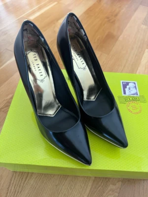 Svarta pumps från Ted Baker - Snygga svarta pumps från Ted Baker storlek 36 med spetsig tå och stilrena gulddetaljer längs sulan och på klacken. Klackhöjd 9-10cm  Endast provade hemma – tyvärr för höga för mig. Försulade direkt efter köp för extra skydd, redo att användas direkt.  Originalkartong finns  Skickas mot frakt eller hämtas på Kungsholmen