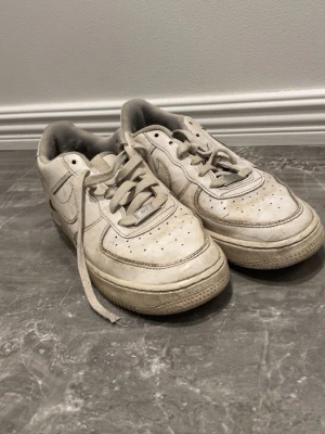 Nike Air Force 1 vita sneakers - Klassiska vita Nike Air Force 1 sneakers med lågt skaft. Skorna har perforerade detaljer på tån, Nike swoosh på sidan och tjock sula med cirkulärt mönster undertill. Tillverkade i läder med textilfoder och snörning framtill.