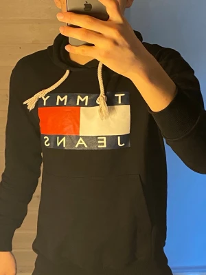 Svart hoodie Tommy Jeans hoodie  - Svart hoodie från Tommy Jeans med stor logga framtill i rött, vitt och blått. Klassisk huva med vita dragsnören och magficka. Mjuk bomullskänsla och ribbade muddar vid ärmslut och nederkant. Perfekt för en chill och stilren look. Står Stl L men det passar s