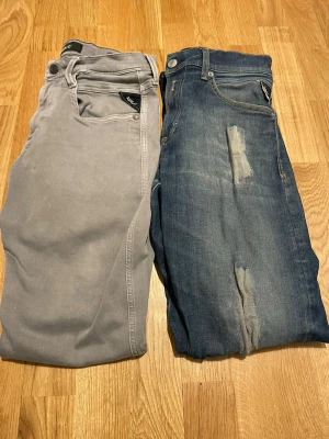 Två par jeans från Replay - Säljer två par jeans från Replay. Ett par är ljusgråa med klassisk femficksdesign och ett par är blå med slitningar och detaljer på benen. Båda har knappstängning och logotyp på fickan. Perfekt för dig som gillar snygga och trendiga jeans.  För mer information skriv gärna. 