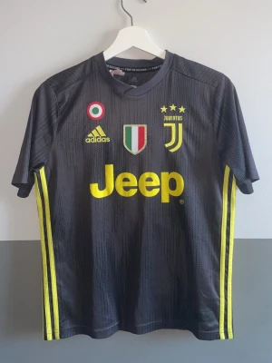 Juventus Ronaldo tröja Adidas - Juventus fotbollströja med Ronaldo och nummer 7 på ryggen. Tröjan är mörkgrå med gula detaljer, Juventus-logga, Adidas-märke och Jeep-tryck på framsidan. Materialet är lätt och andas, perfekt för träning eller match.