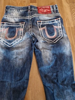 True Religion jeans - Säljer ett par blå bootcut jeans från True Religion med kraftiga kontrastsömmar och stora bakfickor med U-formad brodyr i vitt och rött. Jeansen har slitna detaljer och tvättad look, samt en röd patch bak i midjan. Klassisk femficksmodell i denim.