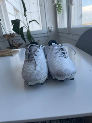 Vita Nike fotbollsskor med guldlogga - Säljer ett par vita Nike fotbollsskor med guldfärgad Swoosh på sidan. Skorna har klassisk snörning, låg profil och dobbar för spel på gräs. Ovandelen är i syntetmaterial med diskreta mönster för bättre bollkontroll. Perfekta för dig som vill sticka ut på planen. Och den är smutsig för att jag har använt den rätt länge men funkar ändå bra. Detta är bra för ni som letar efter några bra fotboll skor som är billiga men inte den bästa.