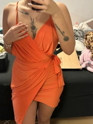 Orange omlottklänning med korsade band - Snygg orange omlottklänning med djup v-ringning och korsade tunna axelband i ryggen. Klänningen har en knytning i sidan och är gjord i ett mjukt, stretchigt material som sitter tight och framhäver figuren. 
