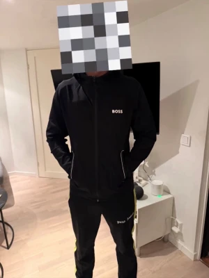 Svart hoodie från BOSS med dragkedja - Svart hoodie från BOSS med vit logotyp på bröstet och dragkedja framtill. Hoodien har huva med snören och två fickor med vita detaljer. Materialet ser ut att vara mjukt och bekvämt, perfekt för en avslappnad stil.