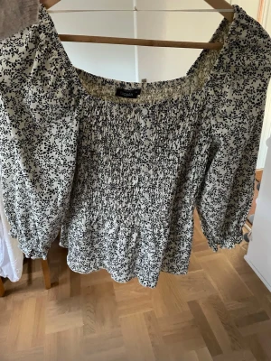 Blommig offshoulder blus från Filippa K - Snygg offshoulder blus från Filippa K med svart blommönster på vit bakgrund. Blusen har smockad överdel och trekvartsärmar med resår vid ärmslut. Perfekt för dig som gillar romantisk och luftig stil. Materialet känns lätt och bekvämt.