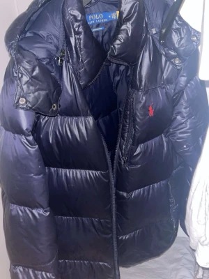 Mörkblå pufferjacka från Polo Ralph Lauren - Snygg mörkblå pufferjacka från Polo Ralph Lauren med huva och dragkedja. Jackan har det klassiska röda Polo-logotrycket på bröstet och breda, vadderade paneler som ger en riktigt puffig look. Perfekt för kalla vinterdagar.