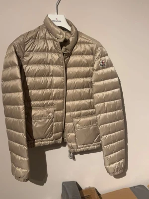 Beige dunjacka från Moncler - Snygg beige dunjacka från Moncler med quiltad design och hög krage. Jackan har två framfickor med lock och tryckknappar samt Moncler-logga på ärmen. Lätt och smidig modell som håller värmen under kalla dagar.