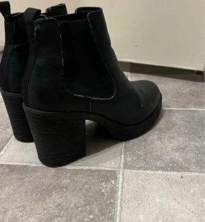 Svarta Chelsea boots med klack - Säljer ett par svarta Chelsea boots med bred klack och elastiska paneler på sidorna. Skorna har grov sula med mönster för extra grepp och är tillverkade i ett matt material som ger en stilren look. Perfekta för dig som vill ha en cool och tidlös stil.
