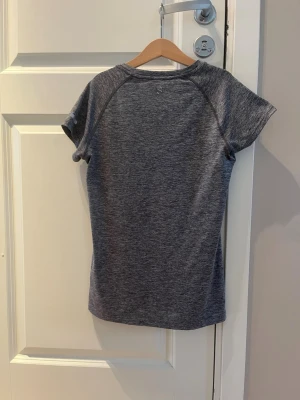 Grå träningströja från H&M - Snygg gråmelerad träningströja från H&M med korta ärmar och rund hals. Tillverkad i ett mjukt och stretchigt funktionsmaterial som andas, perfekt för gymmet eller löprundan. Storleken är exakt 146/152.